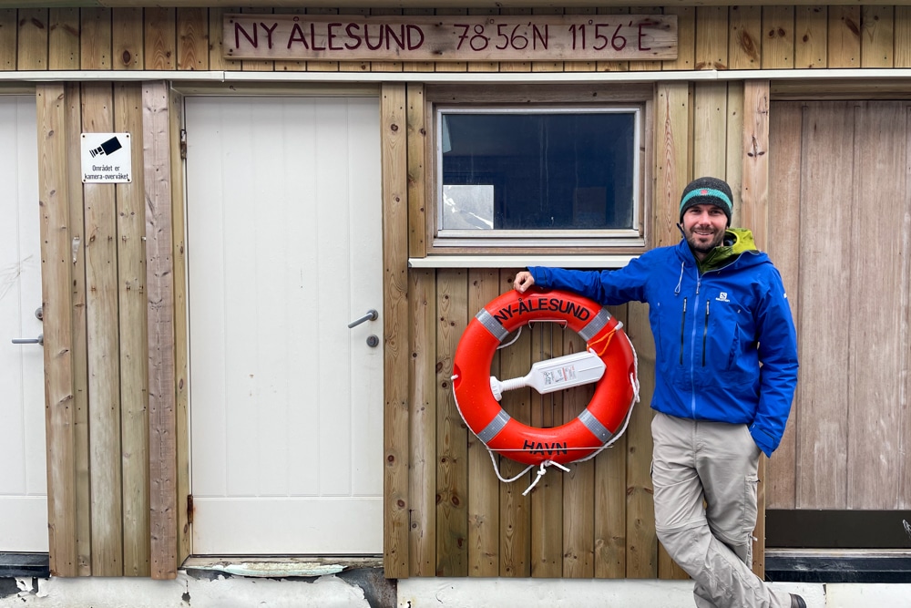 Peter stands in front of a building in Ny-Ålesund with the settlement's coordinates above him