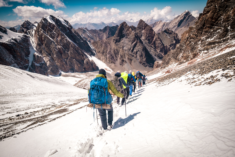 A group of hikers descend a snowy pass using trekking poles