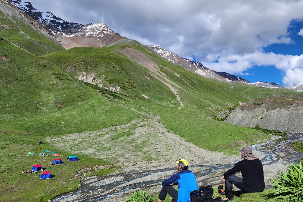 Trekkers relax above a campsite surrounded by mountains and green fields